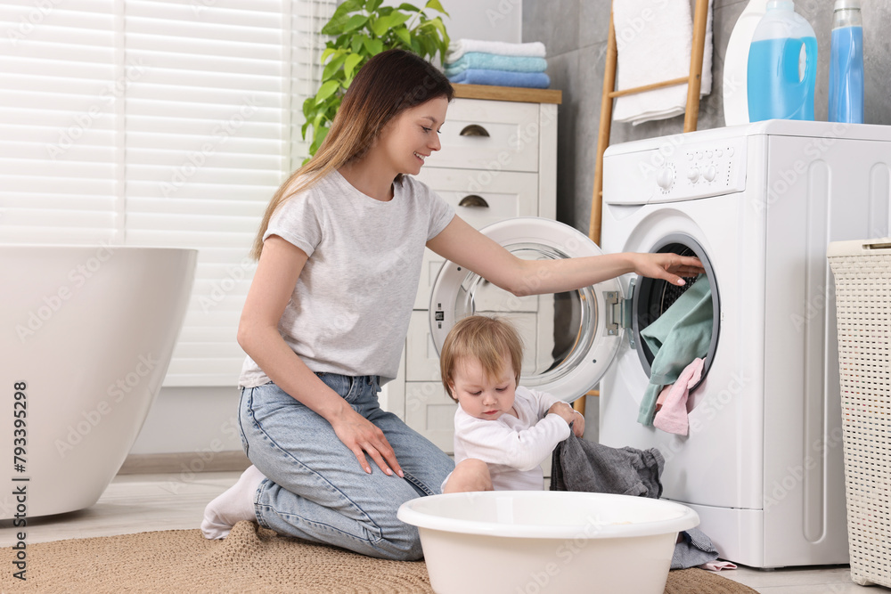 Fototapeta premium Happy mother with her daughter putting baby clothes into washing machine in bathroom
