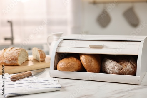 Wooden bread basket with freshly baked loaves and knife on white marble table in kitchen photo