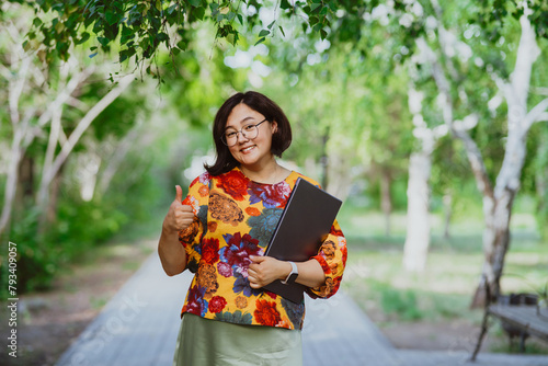 Smiling Asian woman with glasses and a laptop showing thumbs up in a sunny park. Portrait looking at camera. Happy businessman enjoying remote work among green trees outdoors