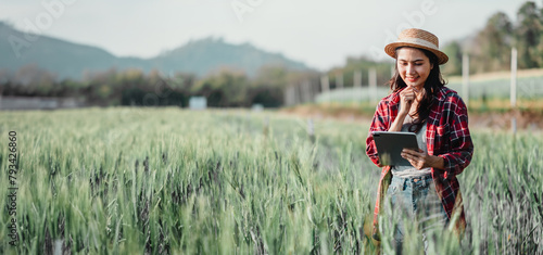 Farmer wearing a straw hat and plaid shirt uses a tablet in a lush wheat field with trees and a house in the background.