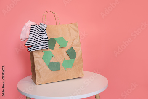 Paper bag with recycling sign with old clothes on table in front of pink background.