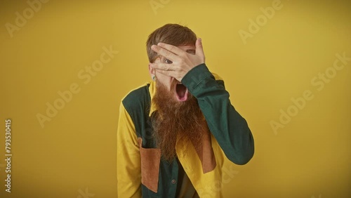 Shy, young redhead guy with glasses, standing over yellow background. he's peeking through his fingers covering face in fear, displaying an embarrassed shock expression.