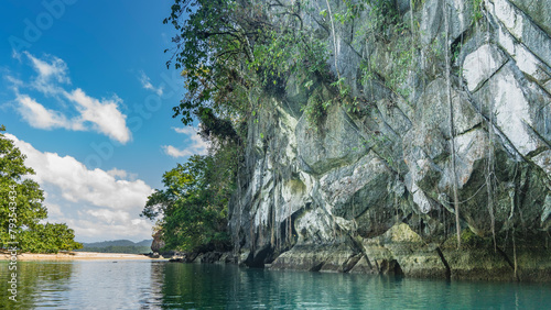 Sheer karst cliffs rise above the emerald calm river. Green vegetation on steep slopes. Glare on the water. Clouds in the blue sky. Philippines. Palawan. Puerto  Princesa. An underground river. photo