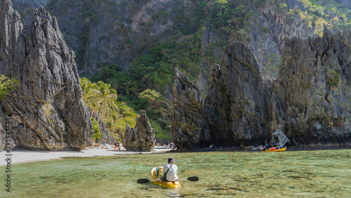 Beautiful karst rocks surround the emerald lagoon. Canoes float in clearwater. People walk along the sandy beach. Green vegetation on steep cliffs. Philippines. Palawan. Bacuit bay. El Nido. 
