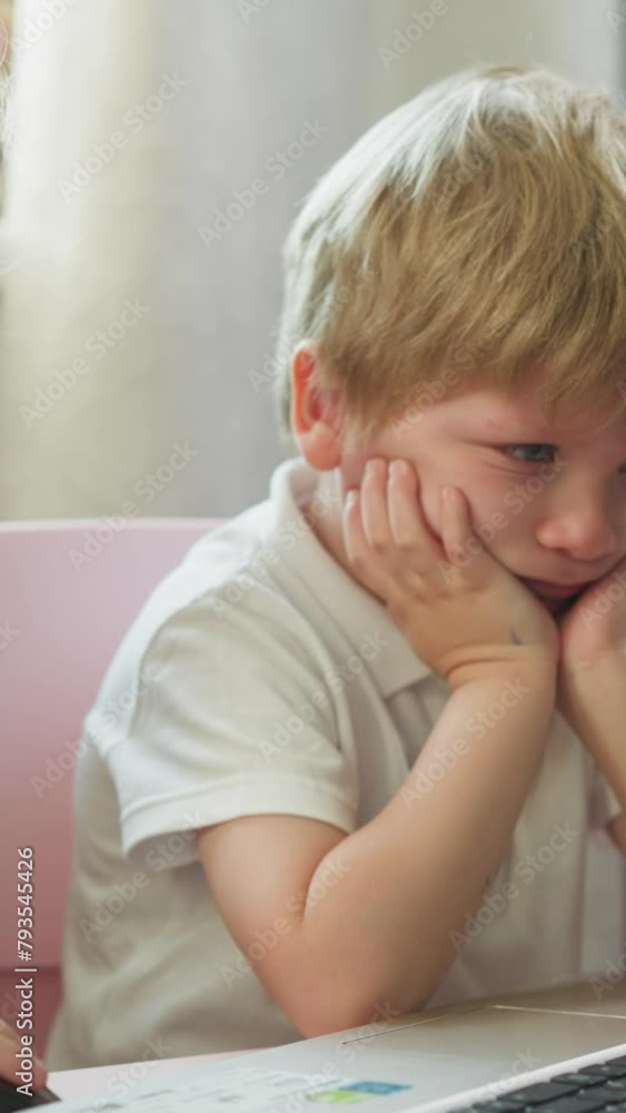 Smiling girl turns on cartoons on laptop to bored brother at table in children room. Little children watch educative videos via computer at home Smiling girl turns on cartoons on laptop to bored brother at table in children room. Little children watch educative videos via computer at home