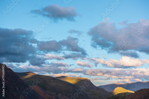 Mountain and hill tops illuminated by sunset light. Rocks silhouettes and gold sunrise colors. Vivid golden sunlight on hills under clouds of sunset tones in blue sky. Shadows of clouds on rocky ridge