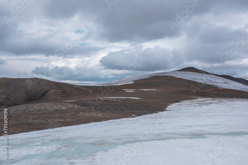 Dramatic view from flat big glacier at high altitude to rocky large mountain top in dome shape under rainy clouds. Atmospheric landscape with rounded stony hill at great height in gray cloudy sky.