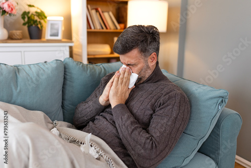 Sick sad man sits on couch at home suffers from runny nose flu disease coronavirus pandemic covid epidemic sneezes. Unwell guy feeling bad fever virus illness symptoms indoor