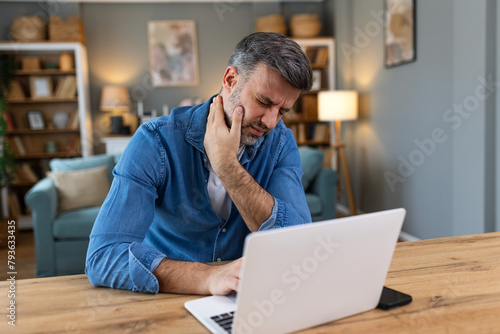 Businessman feeling pain in neck after sitting at the table with laptop. Tired man suffering of office syndrome because of long hours computer work. He is massaging his tense neck muscles