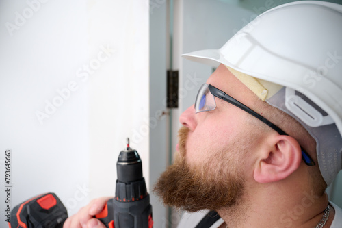Construction worker holding electric cordless screwdriver in hand at construction site. Man worker dressed in work attire and helmet and protective glasses. Home renovation, electric tool for job.