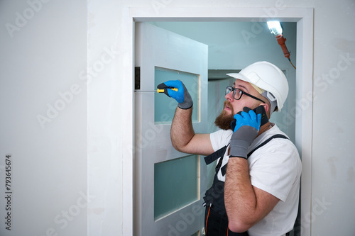 Construction worker with a screwdriver installing a door while speaking on cellphone. Install furniture repair home. Man dressed in work attire, helmet and protective glasses