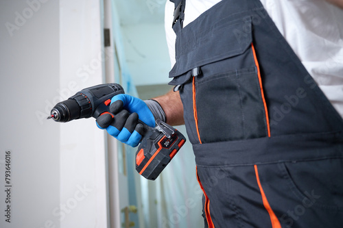 Construction worker holding electric screwdriver in hand at construction site. Accessories for assembling, install furniture, repair home. Man dressed in work attire and helmet and protective glasses.