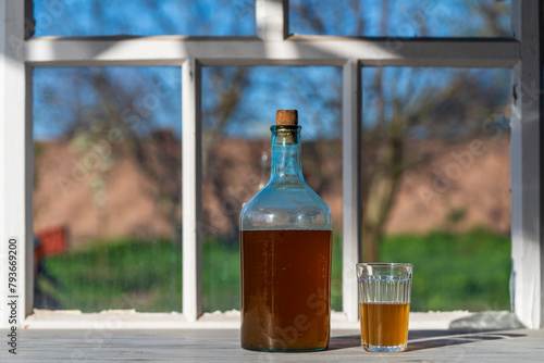 Big bottle with a drink made from fermented birch sap on the windowsill on a warm spring day, closeup. Traditional Ukrainian cold barley drink kvass in a glass jar and glass on table near yard