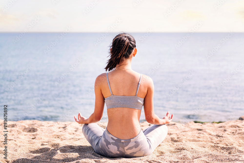 rear view of a young woman doing meditation at beach sitting with legs crossed, concept of mental relaxation and healthy lifestyle