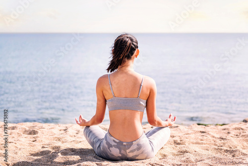 rear view of a young woman doing meditation at beach sitting with legs crossed, concept of mental relaxation and healthy lifestyle