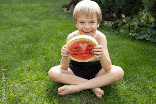 joyful young boy without clothes sits on the grass with a round piece of watermelon, smiling. Summer, good mood, delicious vitamin-rich food