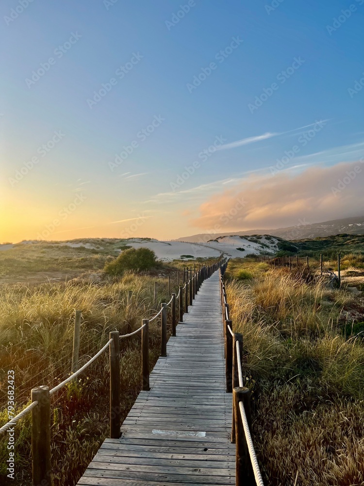 Explore the rustic charm of Portugal's coastline with a wooden bridge at sunset. Bask in the beauty of nature's hues blending seamlessly over sandy shores and tranquil waters