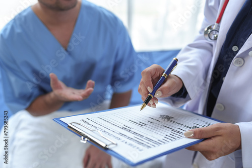 Professional female doctor with stethoscope around her neck talks with male patient, asks patient to fill out form, patient history, disease symptoms, body aches to plan treatment at hospital bed.