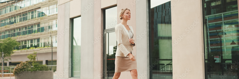 Businesswoman in beige suit walks outside past business building, Panorama