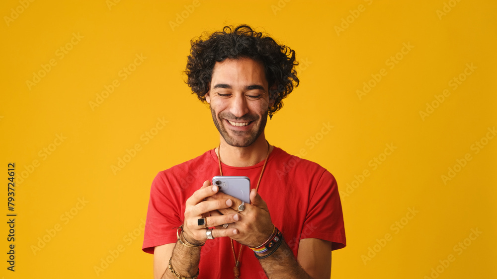 Smiling man with curly hair, dressed in red T-shirt, using mobile phone, typing massages isolated on yellow background in studio Smiling man with curly hair, dressed in red T-shirt, using mobile phone, typing massages isolated on yellow background in studio