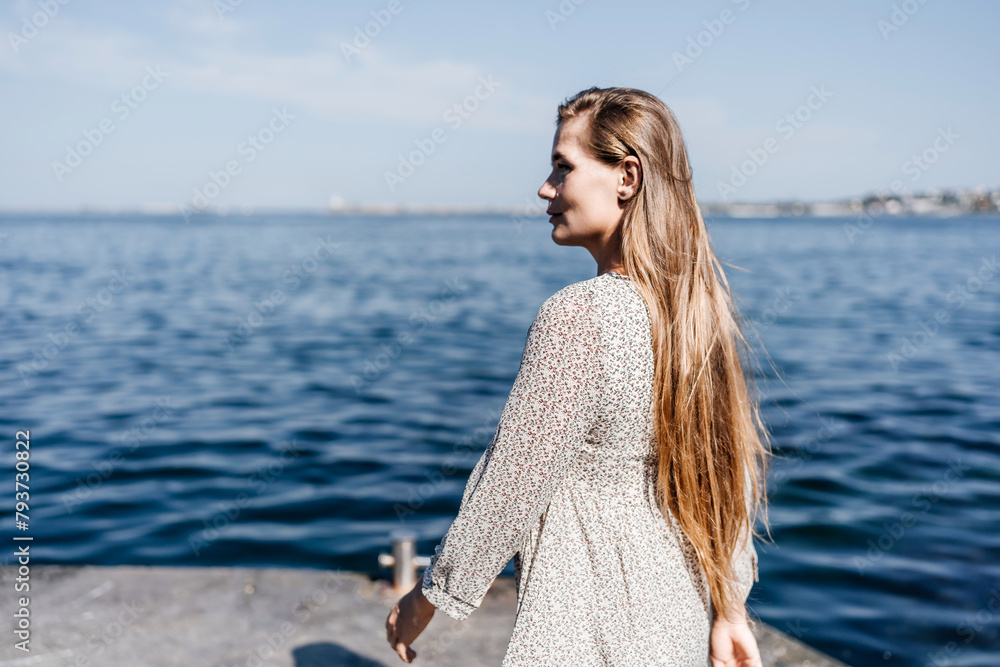 A woman is walking on the beach wearing a dress. The water is calm and the sky is clear.