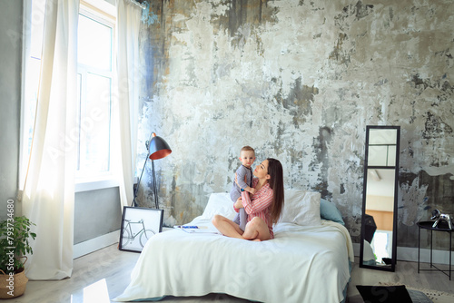 Young millennial mother in pajama sitting on bed in modern light interior, hugging her baby son, playing, lifestyle. Concept of healthy, enjoying motherhood, life. Toddler boy, mom positive emotions photo