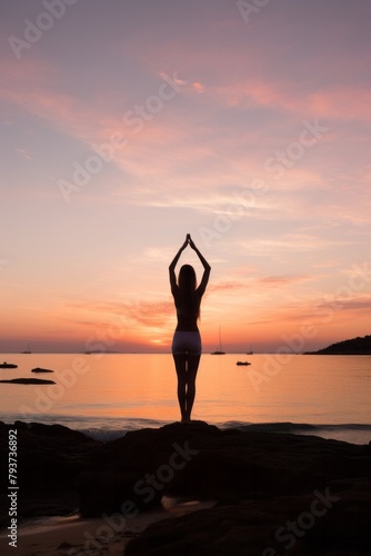 A woman is standing on a beach with her arms raised, practicing yoga at sunrise. The serene beach setting enhances her yoga practice as she connects with nature