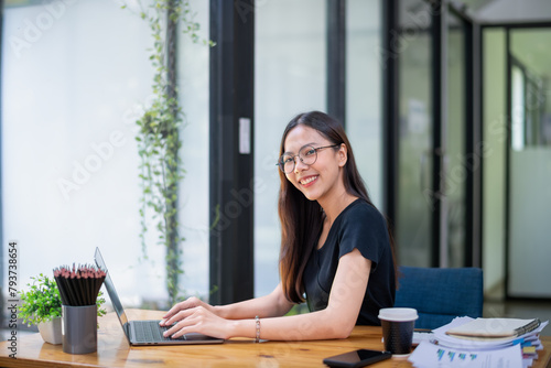 Free Download Cheerful young woman working on her laptop at a wooden table in a bright, plant-filled outdoor setting. - GFXLoad