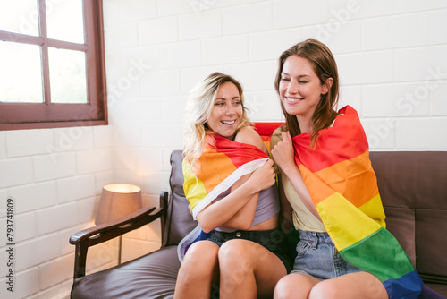 Happy caucasian lesbian couple sitting and embracing together on a sofa covered with Rainbow flags together in the living room, LGBTQ couples spend quality time together.