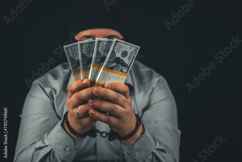 A man in handcuffs and a balaclava mask holds a fan of hundred dollar bills in front of his face on a black background. A man was arrested for a major financial scam.