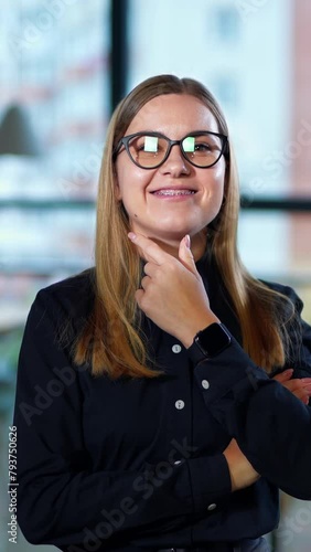 Confident successful young lady in glasses smiling t the camera. Woman standing in office putts of glasses. Office people at the desk at backdrop. Vertical video
