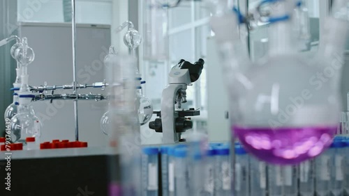 Close-up rack focus no people shot of glass retort with purple chemical fluid on stand in chemistry laboratory, rows of test tubes, microscope in backgroundClose-up rack focus no people shot of glass 