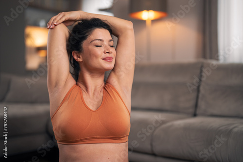 A close-up shot of a contented mid adult woman with closed eyes stretching her arms and exercising at home
