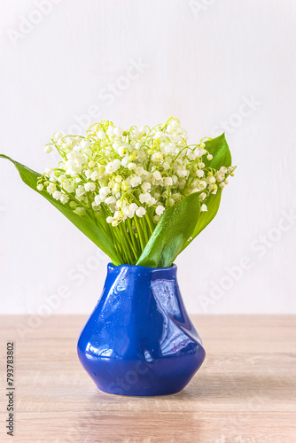Happy May Day greeting card; bouquet of lilies of the valley in blue vase on a wooden table and white background; copy space; vertical picture