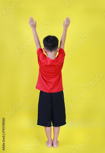 Rear view of Asian little boy child raised hands up. Kid standing and raise arms isolated on yellow background. Image full length.
