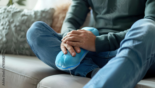 Young man warming his knee with hot water bottle on sofa at home, closeup photo