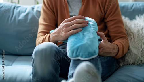 Young man warming his knee with hot water bottle on sofa at home, closeup photo