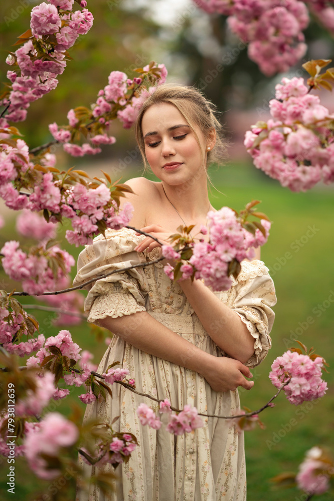 Obraz premium Pretty young blonde girl in vintage lace dress standing in spring park near pink blossom flowers. Tenderness romantic model posing