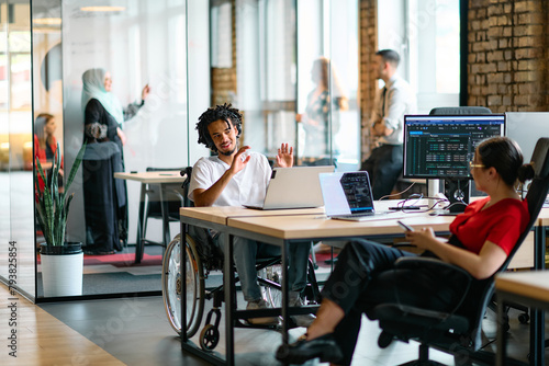 A young business group, including an African American businessman in a wheelchair, collaborates within a modern glass office, actively engaged around a computer and laptop, collectively solving