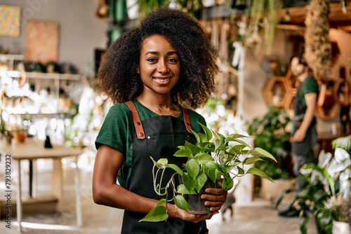 Floristic concept. Portrait of afro young woman in apron standing at modern flower store and holding pot with green domestic flower. Female plant lover transforming favorite hobby into small business. photo