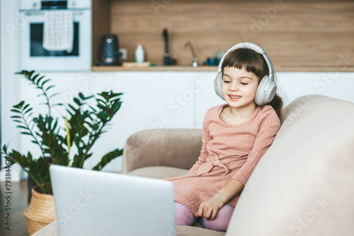 A smiling 5-6 years old girl watching a laptop screen, sitting on a beige couch. Concept: technology-infused relaxation, online education, computer entertainment