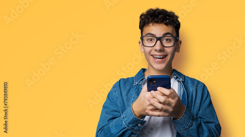 Excited surprised shocked astonished happy curly haired funny young man wear braces glasses spectacles open mouth hold typing cell phone cellular smartphone cellphone isolated yellow background © vgstudio