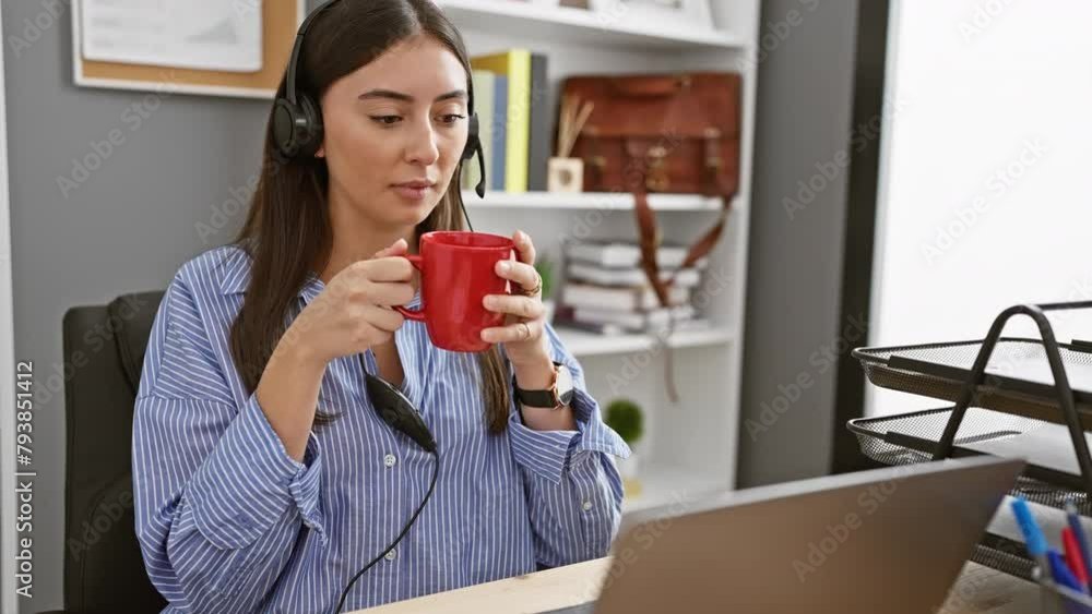 Hispanic woman in headphones holding a red mug at her office desk with a laptop. Hispanic woman in headphones holding a red mug at her office desk with a laptop.