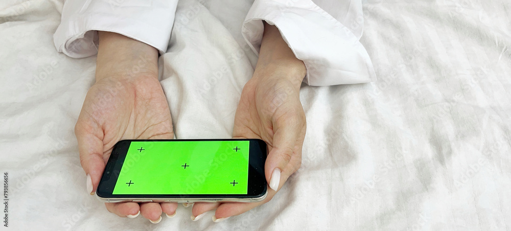 Female hands in a white shirt against the background of a bed hold a phone with a green screen. Mobile Phone Day. Mobile Phone Give Up Day Female hands in a white shirt against the background of a bed hold a phone with a green screen. Mobile Phone Day. Mobile Phone Give Up Day