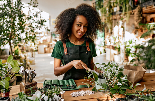Combination of work and hobby. Smiling black woman repotting green flower at flower store using gardening tools, ground and drainage. Qualified gardener taking care of lush plants at modern boutique.