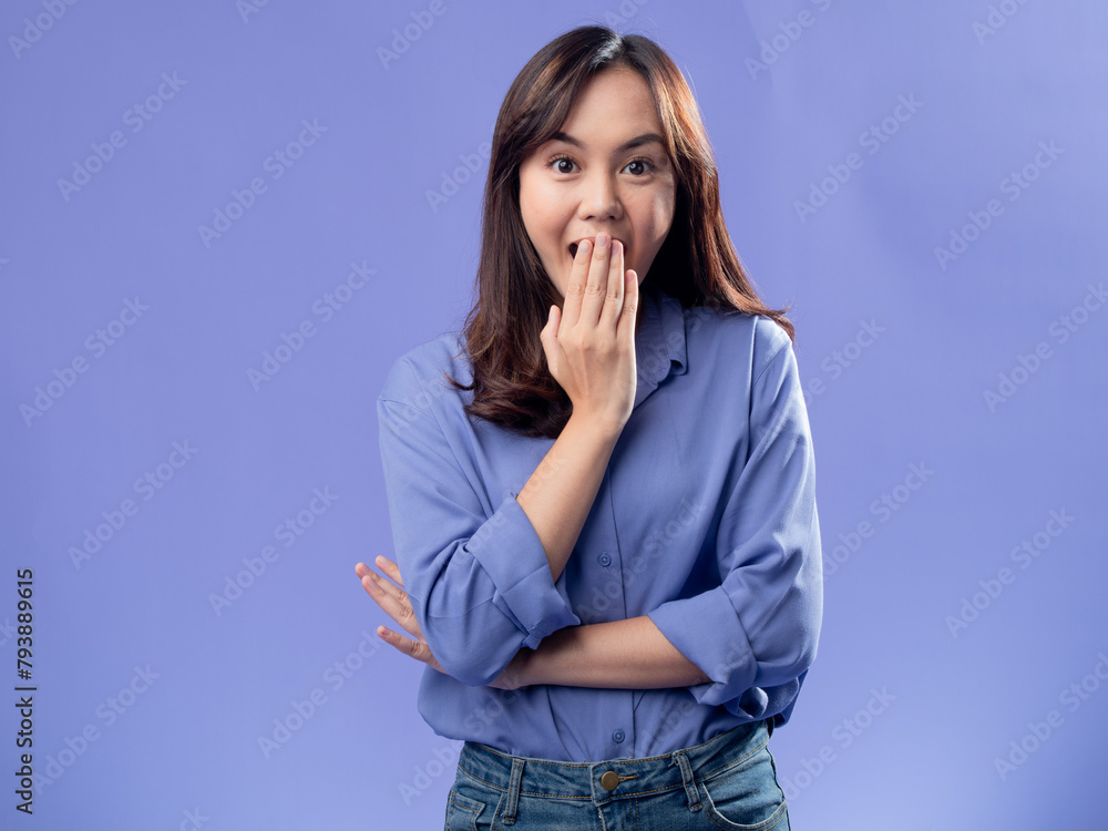 A young Asian woman in a light blue button-up shirt and jeans stands against a solid lavender background, covering her mouth with one hand and widening her eyes, expressing surprise or shock. A young Asian woman in a light blue button-up shirt and jeans stands against a solid lavender background, covering her mouth with one hand and widening her eyes, expressing surprise or shock.