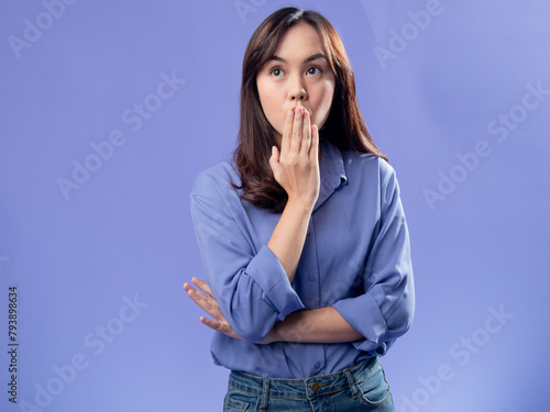 Foto A young Asian woman in a light blue button-up shirt and jeans stands against a solid lavender background, covering her mouth with one hand and widening her eyes, expressing surprise or shock