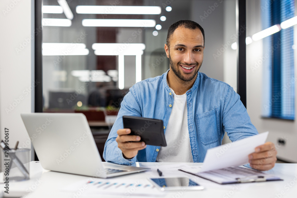 Portrait of a smiling young Hispanic man sitting in a modern office at a desk, working with documents, using a laptop and a calculator