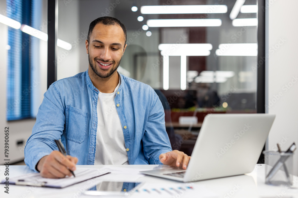 Smiling young hispanic male student sitting in office, working and studying remotely via laptop, making notes in notebook