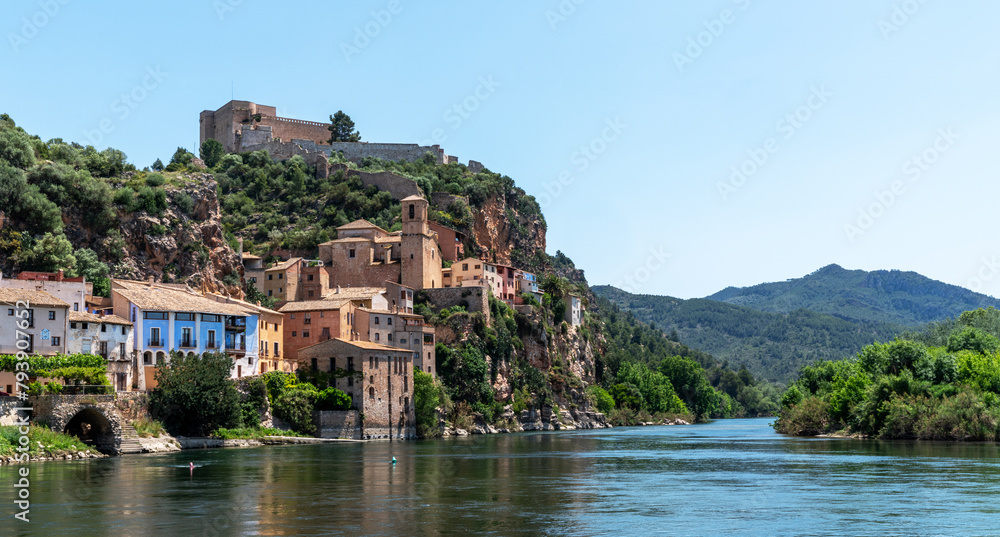 Idyllic riverside village with terracotta-roofed houses nestled on a lush hillside, crowned by a majestic fortress, under the azure sky.